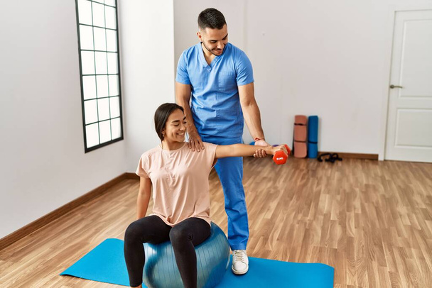 latin man and woman wearing physiotherapist uniform having rehab session using fit ball and dumbbell at rehab center