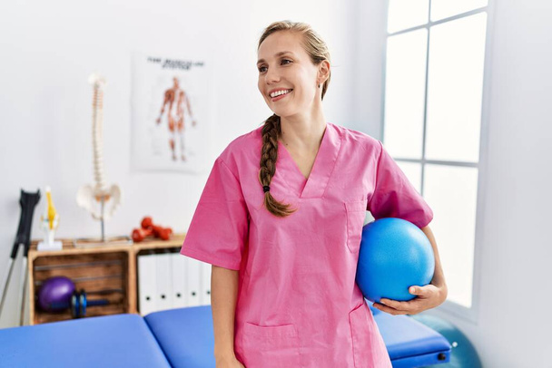 young caucasian woman wearing physiotherapist uniform holding fit ball at physiotherapy clinic